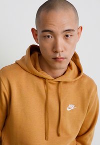 Nike kapuce dzeltenā krāsā, izgatavota no mīkstas auduma. Tam ir priekšējā kabata, kapuce ar šnūru un balta logo detaļa uz krūšu.