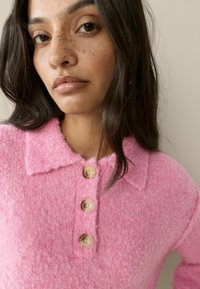 Rožinė megzta palaidinė su apykakle, priekyje su trimis apvaliais mygtukais. Minkšta tekstūra ir šiek tiek puri apdaila.