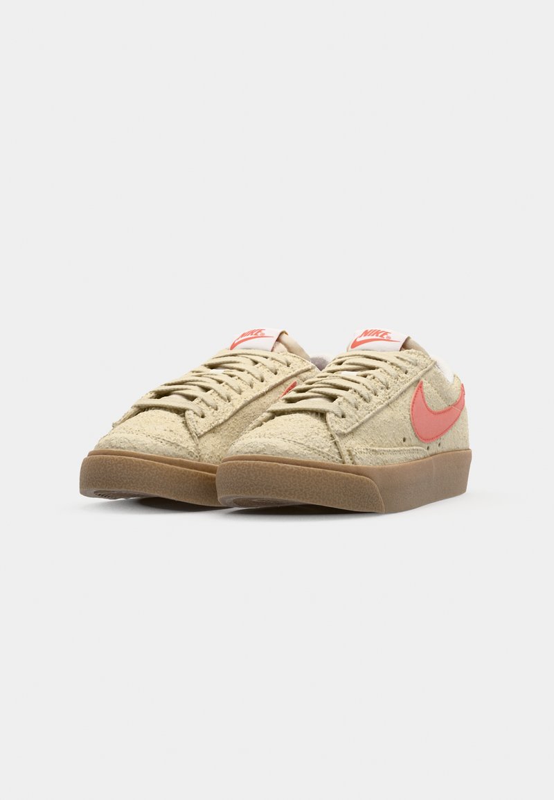 Baskets en suède beige avec un swoosh Nike rose, lacets plats, languette rembourrée et semelle en caoutchouc brun texturé. Design simple et profil bas.