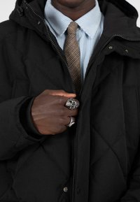 Černá prošívaná bunda s kapucí, nošená přes modrou pruhovanou košili a hnědou kostkovanou kravatou. Stříbrná lebka a ozdobné prsteny na ruce.