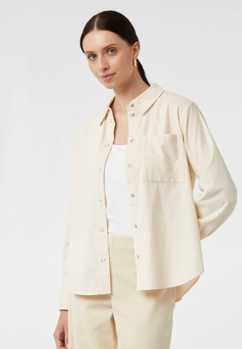 Camicia button-up beige chiaro con colletto, taschino sul petto e hardware argentato. Tessuto morbido con vestibilità rilassata e maniche lunghe.