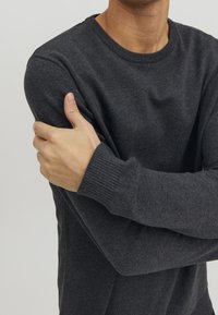 Dunkelgrauer Pullover mit langen Ärmeln und Rundhalsausschnitt. Weiche Textur mit gerippten Bündchen und figurbetonter Passform. Arm über die Brust verschränkt.