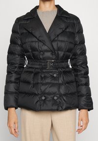 Černá péřová bunda s dvouřadým designem, pásečkem v pase, dvěma velkými knoflíky a lesklým nylonovým materiálem. Texturovaný, prošívaný povrch.