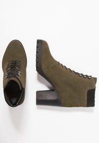 Olijfgroene suède enkellaarsjes met een blokhak, vetersluiting aan de voorkant, metalen oogjes en gestructureerde zwarte accenten op de hak.