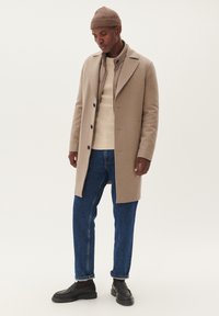 Beige wollen overcoat met grote revers, zwarte knopen, gelaagd over een crèmekleurige geribde sweater, gecombineerd met blauwe jeans en zwarte schoenen.