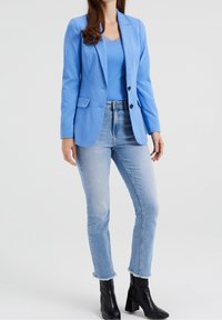 Jasnoniebieska marynarka z dwoma guzikami, zestawiona z pasującym topem na ramiączkach, jasnoniebieskimi dżinsami i czarnymi botkami za kostkę. Gładka tekstura materiału przez cały czas.