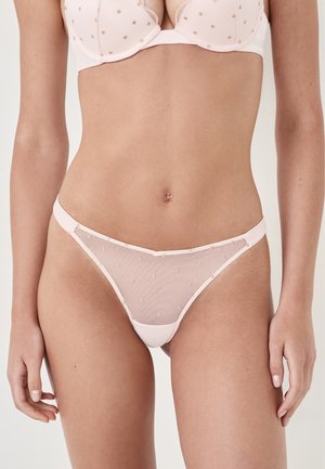 Vrouw in een lichtroze sheer mesh bh en bijpassende slip, staand tegen een egale lichte achtergrond.