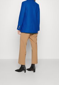 Blazer blu in lana con spalle strutturate, quattro bottoni argentati sui polsini, abbinato a pantaloni beige a vita alta e stivali neri alla caviglia.