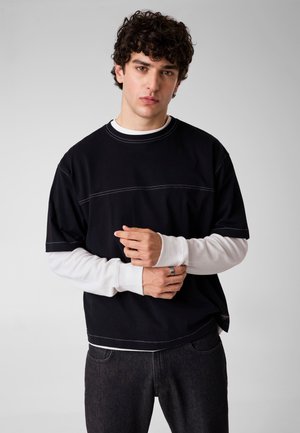 Junger Mann mit lockigem Haar, der ein schwarzes Kurzarmshirt über einem weißen Langarmtop und dunkle Jeans trägt, steht vor einem schlichten Hintergrund.