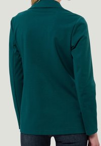 Femme portant une veste à manches longues vert sarcelle avec un col, vue de dos, associée à un jean bleu foncé.