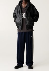 Persona con giacca nera in pelle con zip e cappuccio, camicia scura con numero "91", pantaloni larghi blu navy con strisce bianche sui lati e scarpe nere.