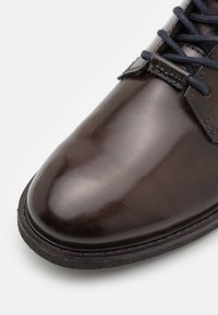 Scarpa in pelle marrone con finitura liscia e lucida; punta arrotondata; lacci blu navy; dettaglio di cucitura lungo la parte superiore e il bordo della suola.