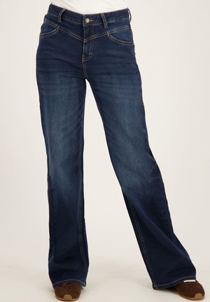 Donkerblauwe spijkerbroek met lichte vervaging, middelhoog taille, rechte pijpen en contrasterende stiksels, gedragen met bruine schoenen.