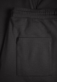 Primer plano de una cinturilla elástica negra con un gran bolsillo cuadrado cosido en una tela suave, que presenta un sutil cosido texturizado y un pequeño logo bordado.