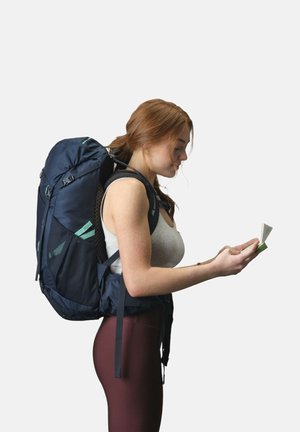 Marineblauer Rucksack mit einer strukturierten Netzrückseite, verstellbaren Trägern und hellgrünen Akzenten. Der Benutzer hält eine Karte und trägt weinrote Leggings.