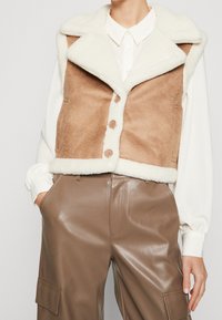 Brun faux shearling-väst med krämfärgad krage och detaljer, med knäppning fram, sidofickor, bärs över en vit skjorta och bruna läderbyxor.