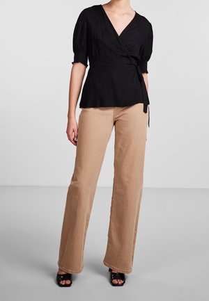 Donna che indossa una blusa nera avvolgente con maniche a sbuffo, pantaloni beige a gamba larga e sandali neri con tacco e punta aperta, su uno sfondo semplice.