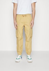 Khaaki cargobyxor med elastiska manchetter, flera sidofickor och dragsko i midjan; kombinerat med vita sneakers och en vit t-shirt.