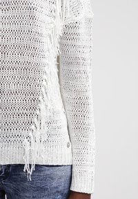 Weißer Strickpullover mit lockerer Webart, mit fransigen Kanten und rundem Halsausschnitt. Beinhaltet ein kleines Knopfdetail am Saum.
