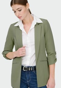 Blazer vert olive à manches trois-quarts, deux poches avant et une ceinture noire sur un jean bleu ; chemise blanche à col en dessous.