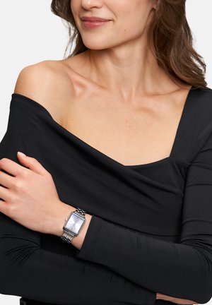 Silver metal watch with rectangular face and linked band worn on a wrist, paired with a black off-shoulder top, showing smooth fabric texture.