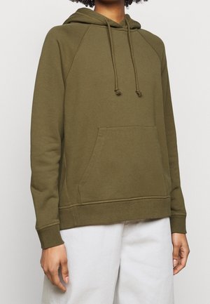 Person wearing olive green hooded sweatshirt with front pocket and drawstrings, paired with light-colored pants against a plain background.