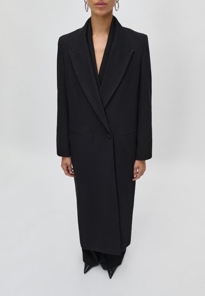 Donna che indossa un lungo cappotto nero doppiopetto con ampie paramonture, sopra pantaloni neri e scarpe nere a punta, in piedi davanti a uno sfondo semplice.