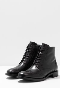 Botas de cuero negras hasta el tobillo con un acabado suave, parte delantera con cordones, detalles de brogue y un tacón bajo apilado.