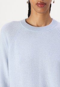 Suéter azul claro de tejido suave, con cuello redondo y mangas raglán, con una textura lisa y un detallado minimalista.
