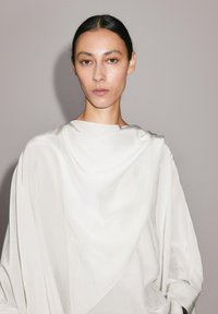 Vêtement en tissu drapé blanc et élégant avec une texture douce, présentant un col rond et une silhouette fluide, porté par un mannequin aux cheveux foncés.