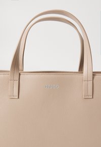 Beige lær tote bag med teksturert overflate, to håndtak øverst, åpen topp-design og en sølvfarget logo tydelig vist på den ene siden.