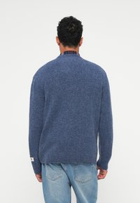 Maglione blu lavorato a maglia con una finitura testurizzata e un taglio rilassato. Presenta maniche lunghe e un'etichetta sul lato sinistro inferiore, indossato sopra una camicia a quadri.