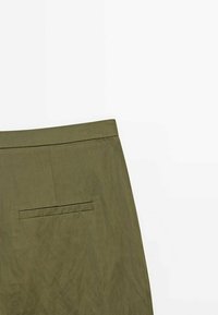 Tissu vert olive avec une poche fendue horizontale, montrant une partie du dos d'un vêtement sur un fond blanc uni.