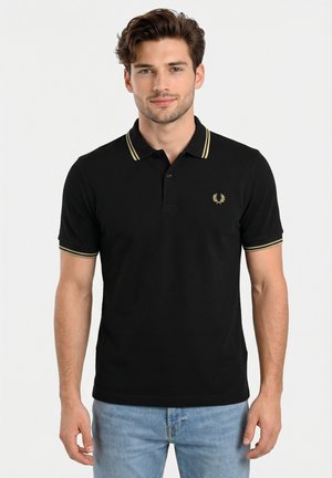 Hombre joven con camiseta polo negra con ribete dorado en el cuello y las mangas, combinado con pantalones vaqueros azul claro, posando frente a un fondo liso.