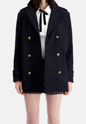 Kobieta w czarnym dwurzędowym płaszczu ze złotymi guzikami, na białej bluzce z czarną wstążką i czarno-białych szortach w wzory.