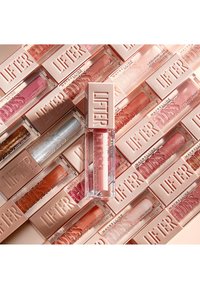 Gloss à lèvres transparent dans un tube rectangulaire avec une accent doré. Emballage environnant dans différentes nuances de rose et de nude, toutes avec le texte "LIFTER GLOSS".