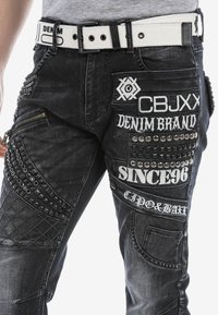 Zwarte denim jeans met verschillende texturen, geborduurde logo's, decoratieve studs en een witte riem met een zwarte gesp.