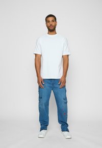 Hvid bomulds t-shirt med korte ærmer, parret med blå cargo jeans med flere lommer og et logo patch. Hvide sneakers fuldender looket.
