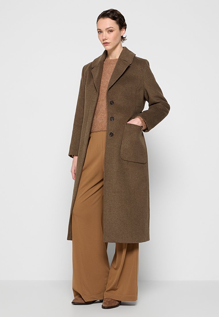 Langer, brauner Wollmischungsmantel mit Reverskragen, drei Knöpfen und Seitentaschen, kombiniert mit einem braunen Pullover und weiten Hosen.