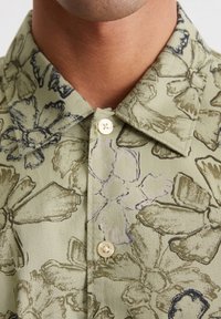 Groene bloemen knoopshirt heeft een kraag, licht materiaal met grote bloemenprints in donkere tinten, en een ronde, lichtgekleurde knop.