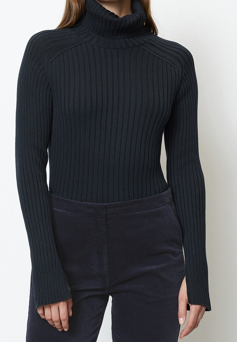 Musta, ribbineulottu pooloneule pitkillä hihoilla ja istuvalla mallilla, esiteltynä tummien housujen kanssa, korostaen tekstuuria ja neulekuvioita.