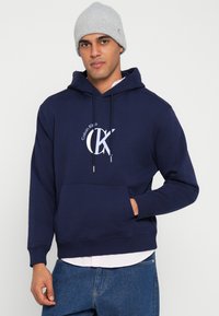 Tmavomodrá mikina s kapucňou z bavlneného zmesi, s predným vreckom, potlačou loga na hrudi a sťahovacou šnúrkou na kapucni. Kombinovaná so svetlými rifľovými nohavicami.