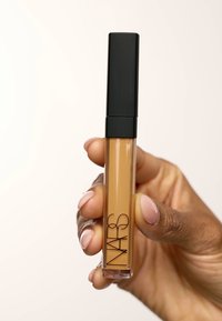 NARS concealer i en klar tube med svart hette, som viser en tan væske inni. Produktet holdes i en hånd med lys hudfarge.