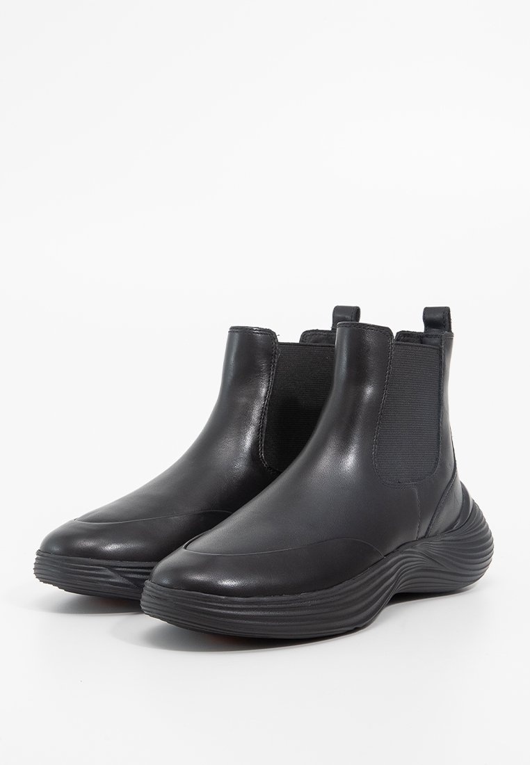 Une paire de bottines en cuir noir avec des panneaux latéraux élastiques et des semelles en caoutchouc épaisses et nervurées, présentée sur un fond blanc.