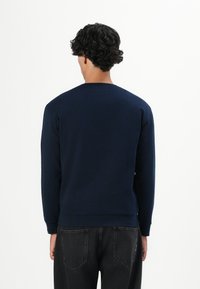 Hombre con cabello rizado negro que lleva una camisa de manga larga azul marino y jeans negros, de pie de espaldas contra un fondo blanco.