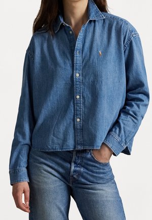 Persoon draagt een ontspannen vallend blauw denim overhemd met knopen en blauw spijkerbroek met medium-wassing, met één hand in de zak tegen een effen achtergrond.