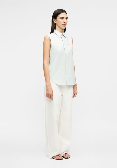 Femme portant une chemise sans manches bleu clair à boutons, un large pantalon blanc et des tongs noires, debout devant un fond blanc uni.