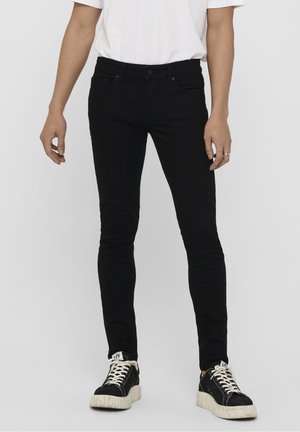 Mann trägt schlanke schwarze Jeans, weißes T-Shirt und schwarze Turnschuhe mit weißen Sohlen und Schnürsenkeln, steht vor einem einfarbigen Hintergrund.