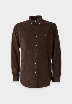 Jeune homme debout portant une chemise marron à manches longues boutonnée, un jean bleu clair à jambes larges et des baskets blanches sur fond blanc.