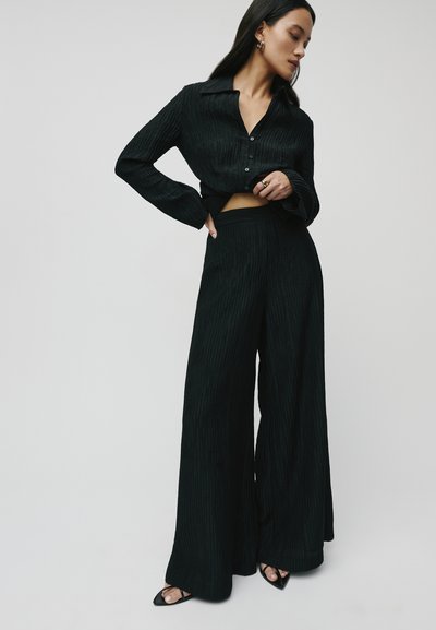 Zwart twee-delig outfit bestaande uit een gekraagde, cropped long-sleeve shirt en wijde broek met een gestructureerd ribbelpatroon. Zwarte haksandalen.